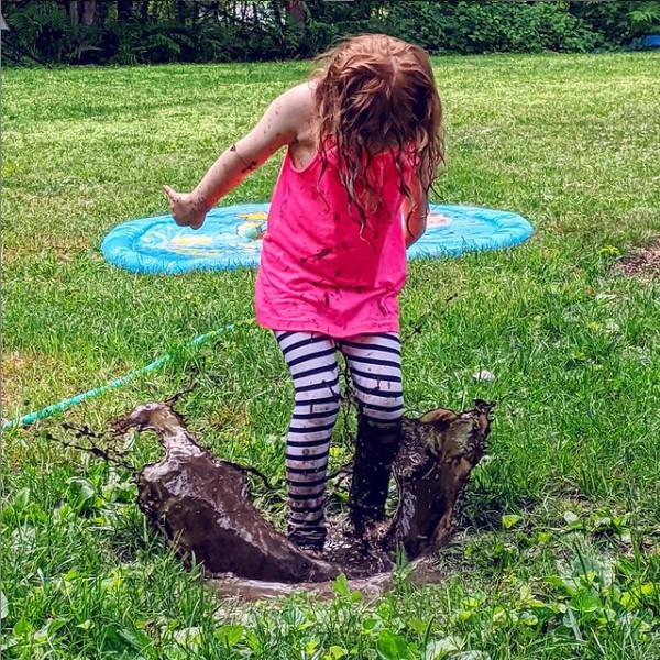 A child with long hair wearing a bright pink shirt with white and black striped leggings jumping in a mud puddle, creating a giant splash. There is a blue splash pad and a garden hose on grass in the background.