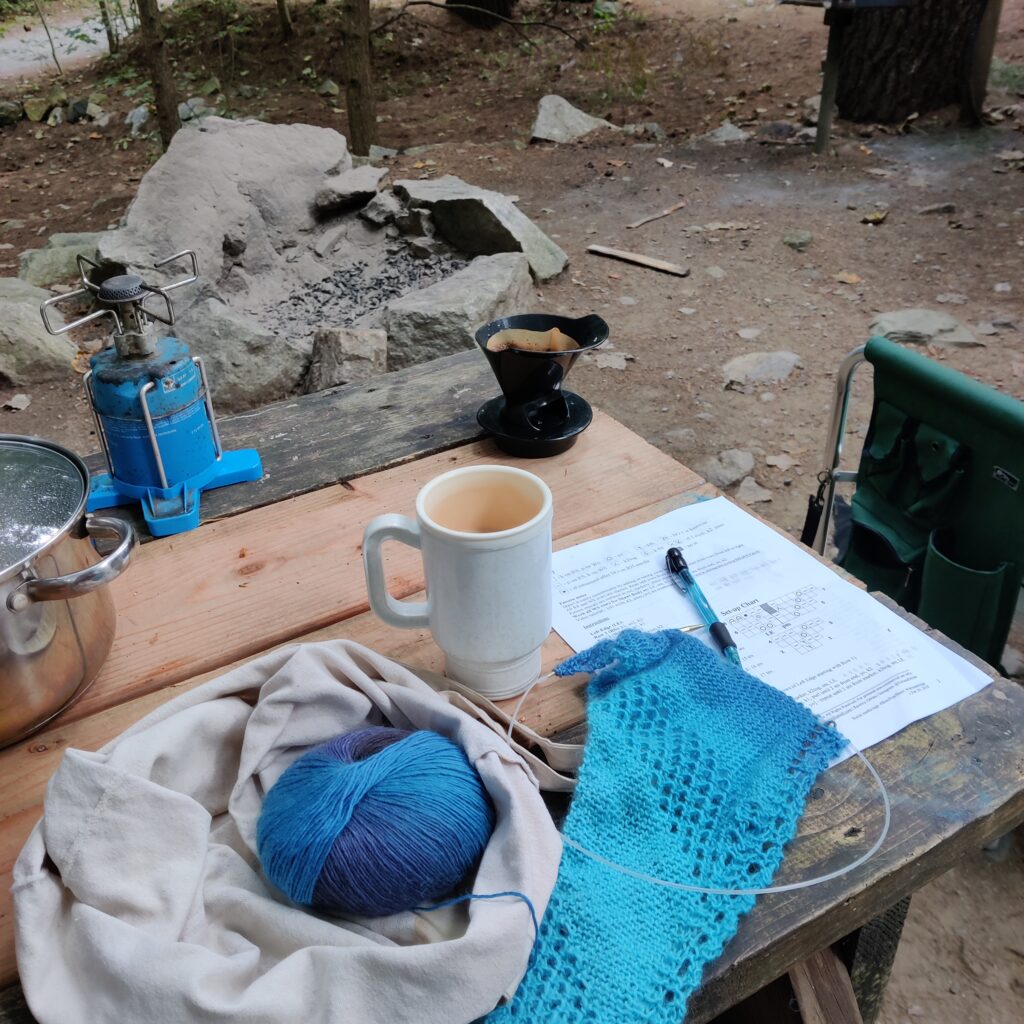 A knitting project in progress using a yarn of varying blue shades sitting on a picnic table outside. There is a burner, a coffee mug, a pot, and a coffee drip on the table as well. There is  a stone fire pit in the background. 