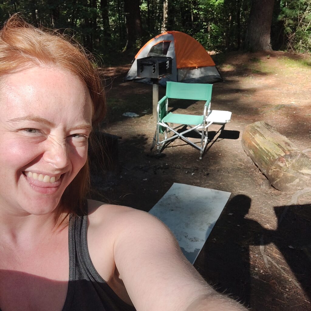 Me smiling in front of my camping setup, including my tent, a folding chair with table, and a very dirty yoga mat.