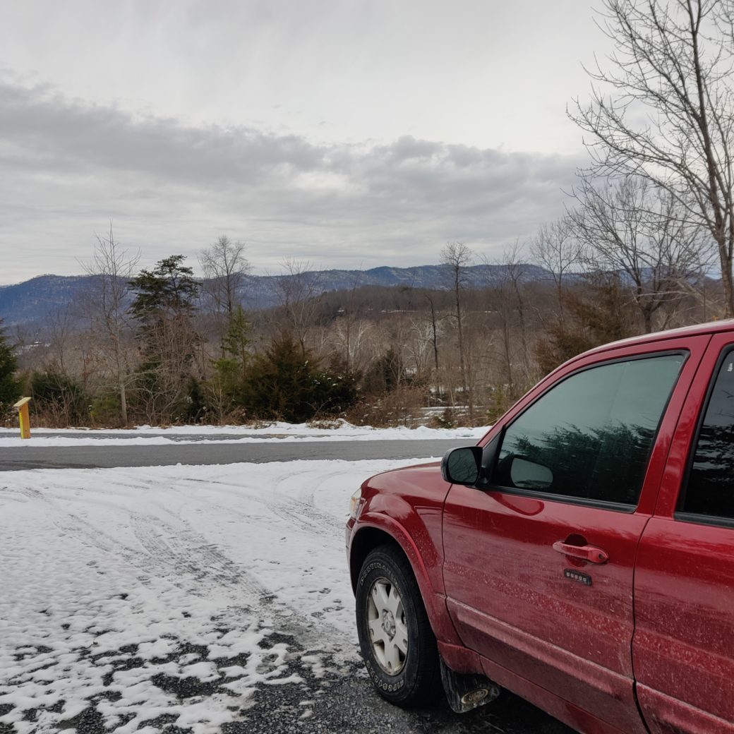 My big red car parked in a snowy area overlooking a mountain view.