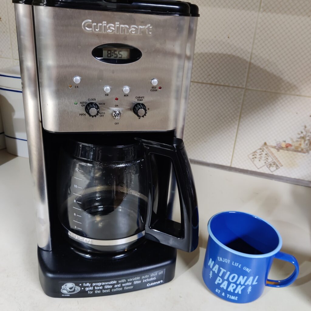 A blue mug with light blue text reading "enjoy life one National Park at a time" sitting beside a coffee maker