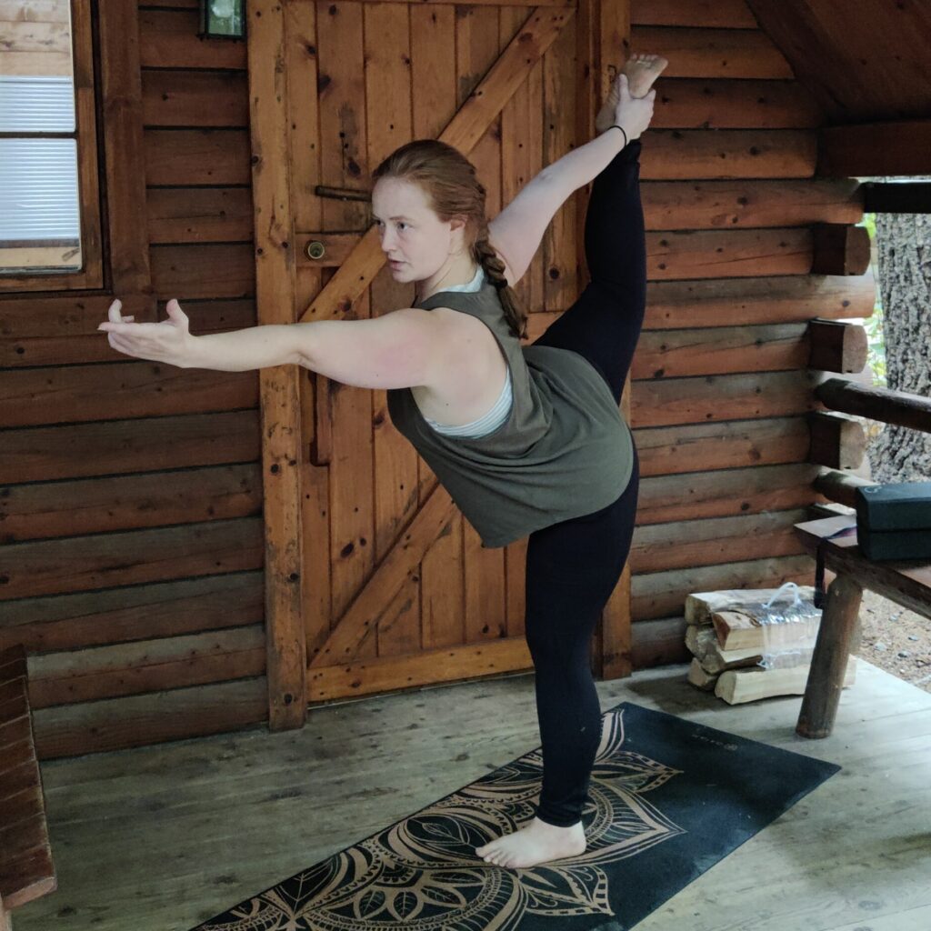 Halliday practicing yoga in standing bow pose. Her hair is in a braid and she is on the porch of a log cabin.